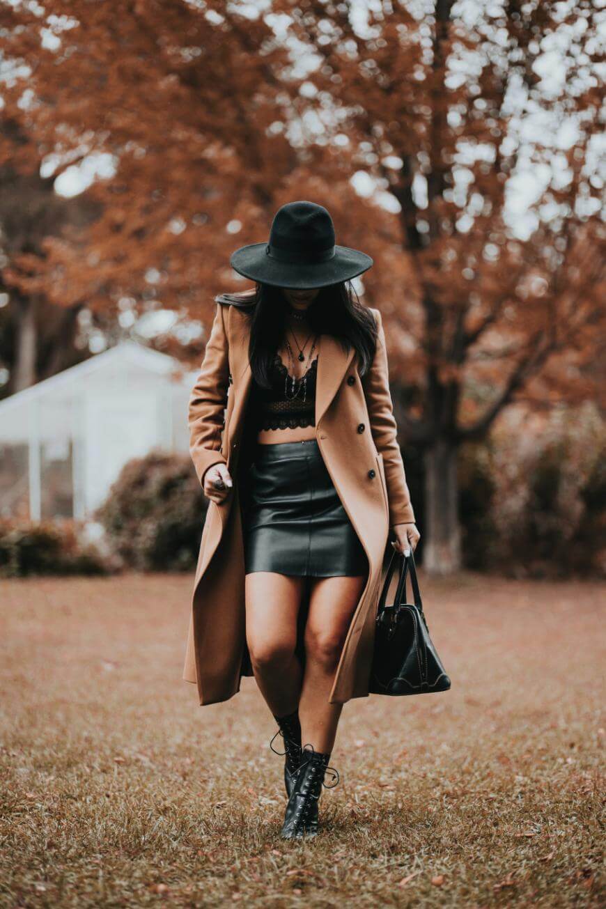 A woman wearing a tan coat, black hat, black lace top, black skirt, and black boots walks on grass with autumn trees in the background, holding a black handbag.