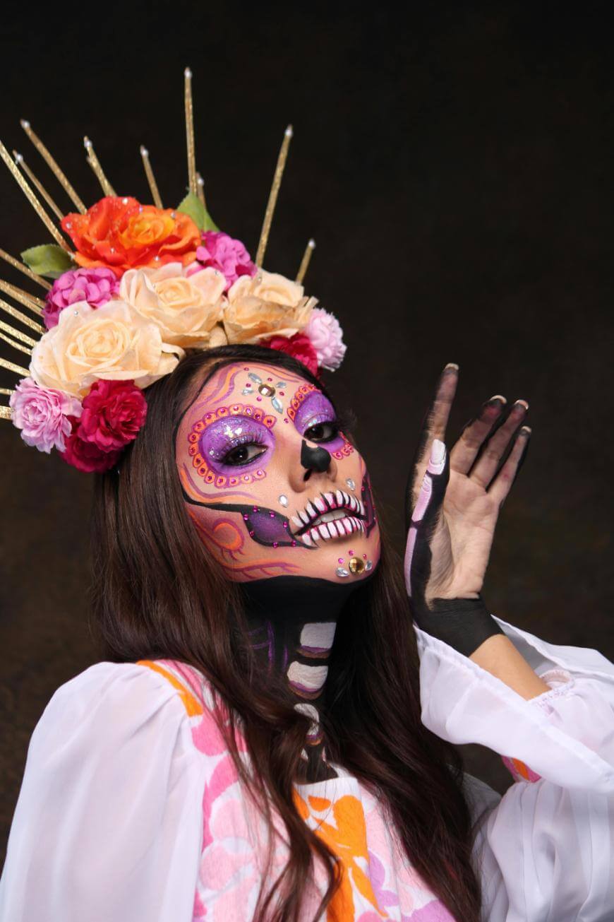 A person dressed for Día de los Muertos with colorful sugar skull face paint, a crown of vibrant flowers, and a white traditional dress with pink and orange patterns, posing with one hand raised.