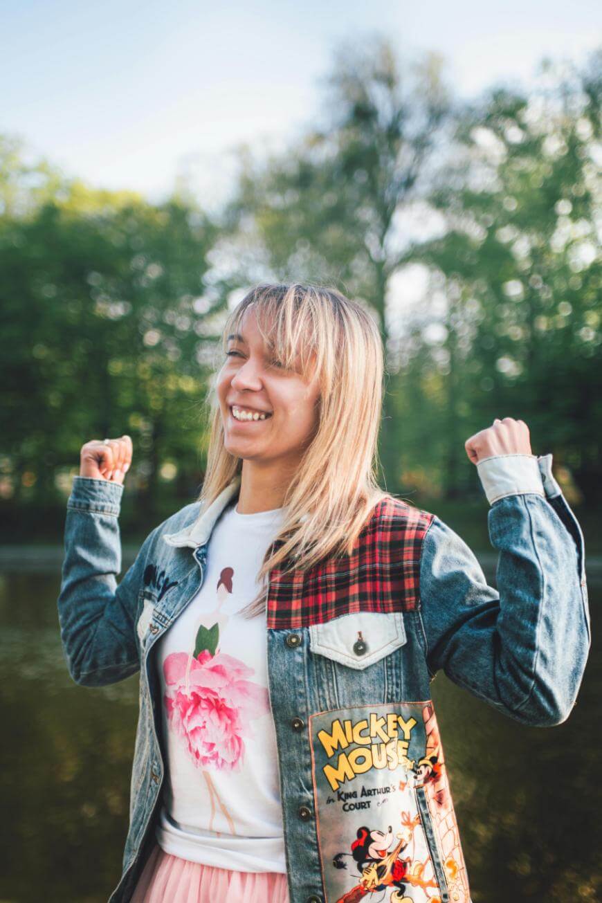 A smiling woman stands outdoors with her arms raised in a joyful pose. She wears a denim jacket with Mickey Mouse patches and a floral shirt, with trees and a pond in the background.