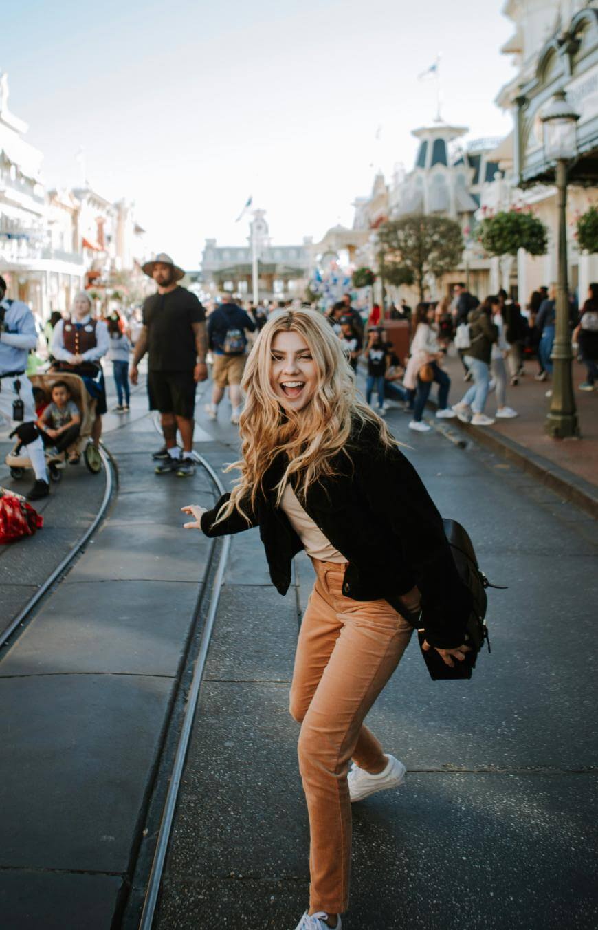 A young woman with long blonde hair, wearing a black jacket and tan pants, smiles and poses playfully on a busy street lined with people and shops on a sunny day.