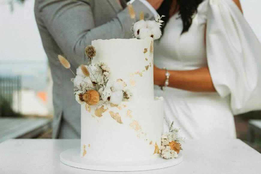 A white two-tier wedding cake decorated with dried flowers and gold accents sits on a table; a bride in a white dress and a groom in a gray suit stand behind it, partially visible.