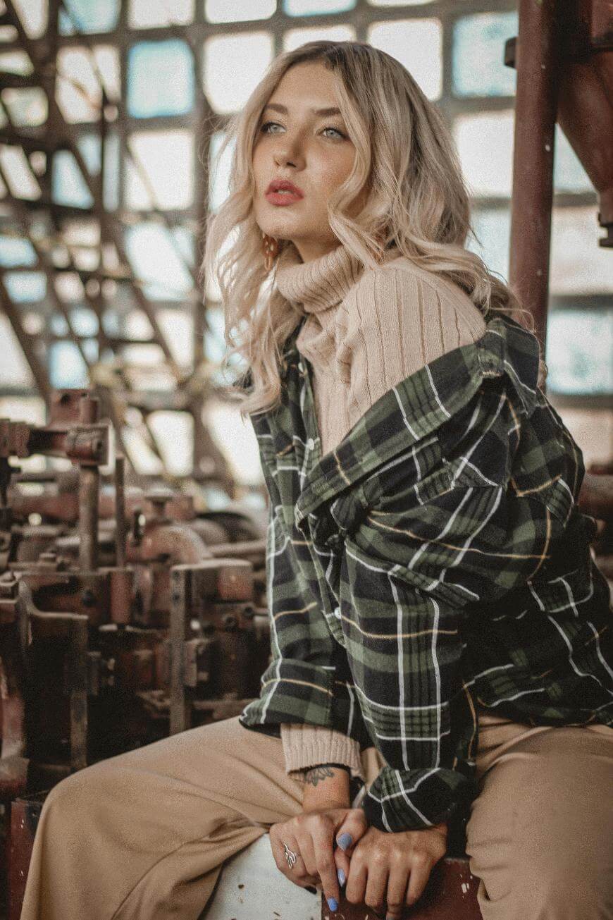 A woman with wavy blonde hair wears a cream turtleneck sweater under a green plaid shirt and beige pants, sitting in an industrial setting with metal structures and large windows in the background.