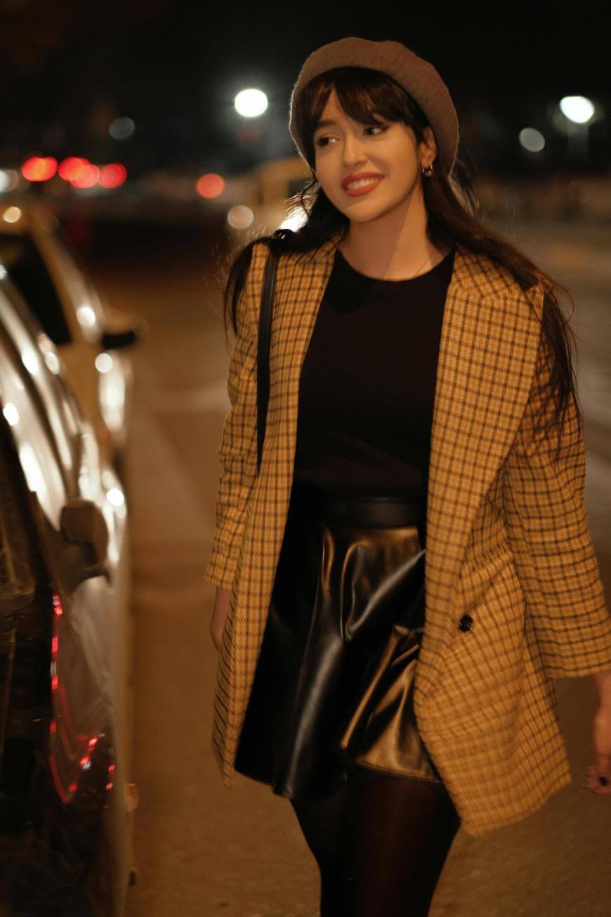 A woman wearing a plaid coat, black skirt, and beret walks along a street at night, smiling. Blurred lights and cars are visible in the background.