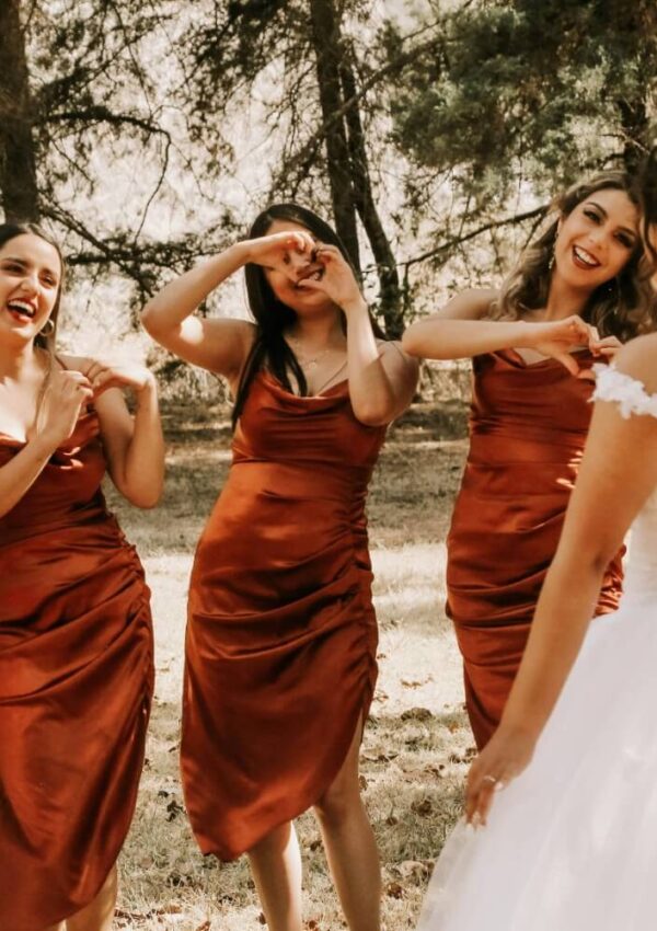 Three bridesmaids in matching rust-colored dresses smile and make heart shapes with their hands toward a bride in a white gown, outdoors among trees in natural light.