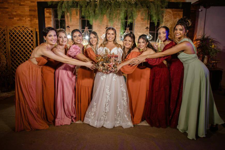 A bride in a white dress stands smiling with eight bridesmaids in colorful dresses, all reaching toward her bouquet and posing happily together indoors against a decorated brick wall.