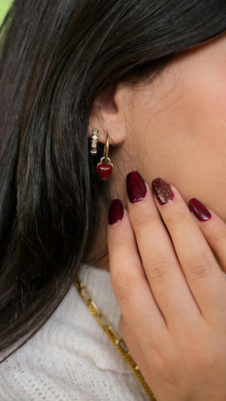 A close-up of a womans ear wearing a gold hoop earring with a red heart charm. Her hand, with deep burgundy and glittery gold nail polish, touches her face. She has long dark hair and wears a white sweater.