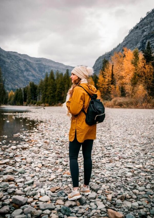 A woman wearing a yellow jacket, black leggings, and a beanie stands on a rocky riverbank with a black backpack. Autumn trees and mountains surround her under a cloudy sky.