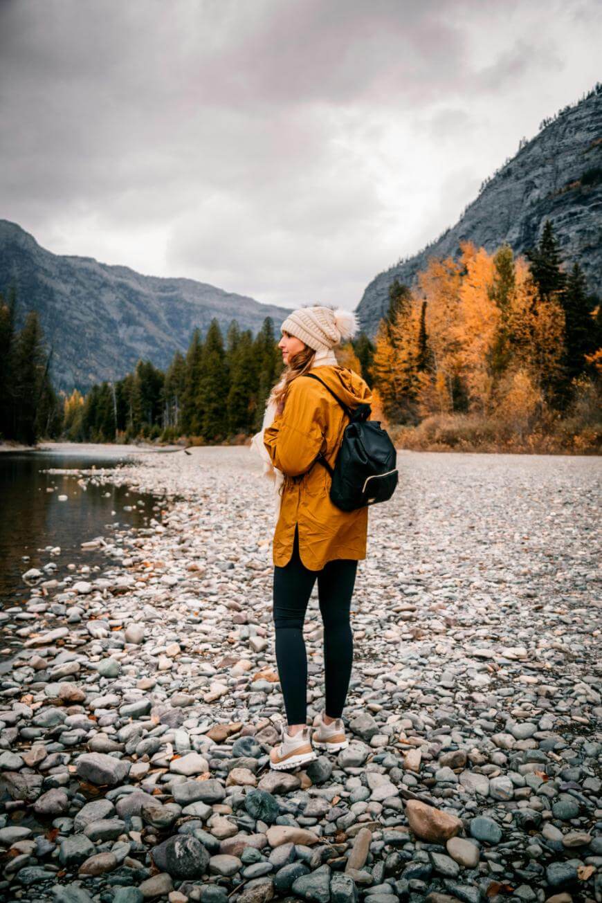 A person in a yellow jacket, black leggings, and a beanie stands on a rocky riverbank, wearing a backpack. Autumn trees and mountains are in the background under a cloudy sky.