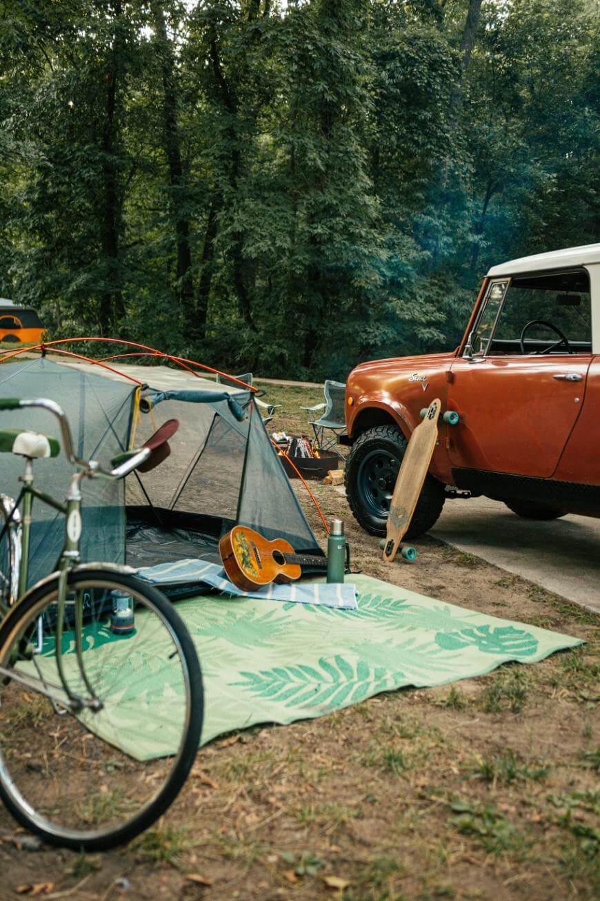 A campsite with a pitched tent, a bicycle, a guitar, a thermos on a green mat, a skateboard, and an orange vintage car parked nearby, surrounded by trees in a forested area.