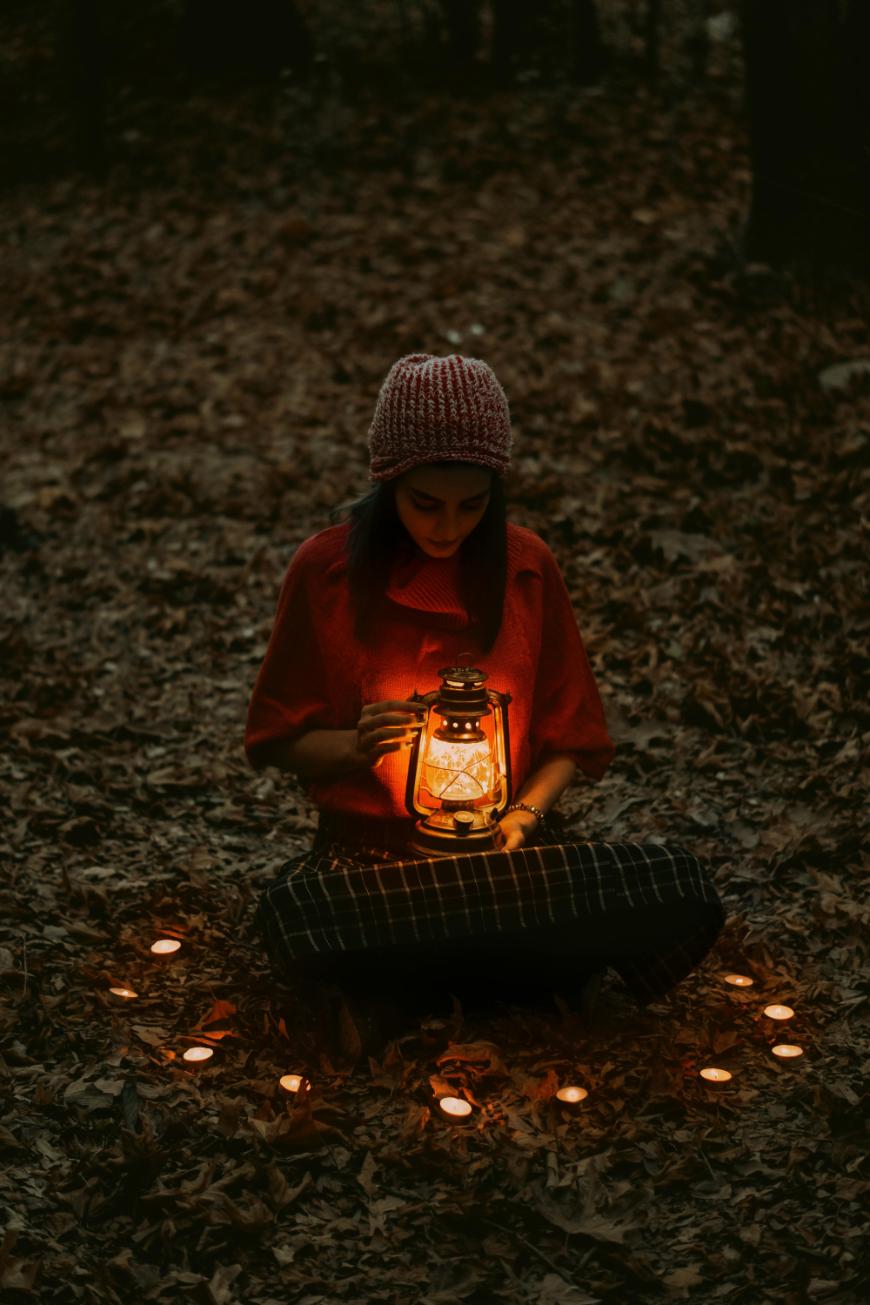 A person in a red sweater and knitted hat sits cross-legged on autumn leaves at night, holding a glowing lantern, surrounded by small lit candles arranged in a circle.