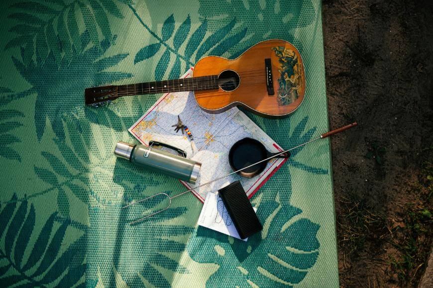 A guitar, a thermos, a notebook, a pen, and a small portable speaker are arranged on a green mat with leaf patterns, hinting at an outdoor setting. A city map lies open beneath the items.