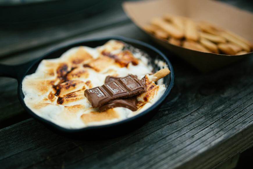 A small cast iron skillet filled with toasted marshmallows, melted chocolate, and a biscuit on top, sits on a wooden surface. In the background, a paper tray holds more biscuit sticks.