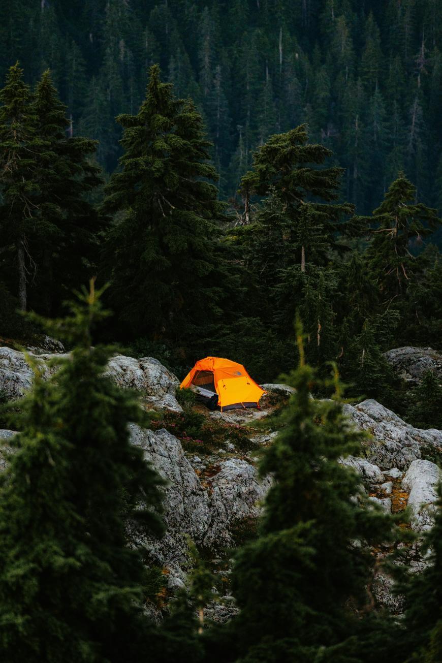 A bright orange tent is set up on rocky ground surrounded by dense evergreen trees in a mountain forest.
