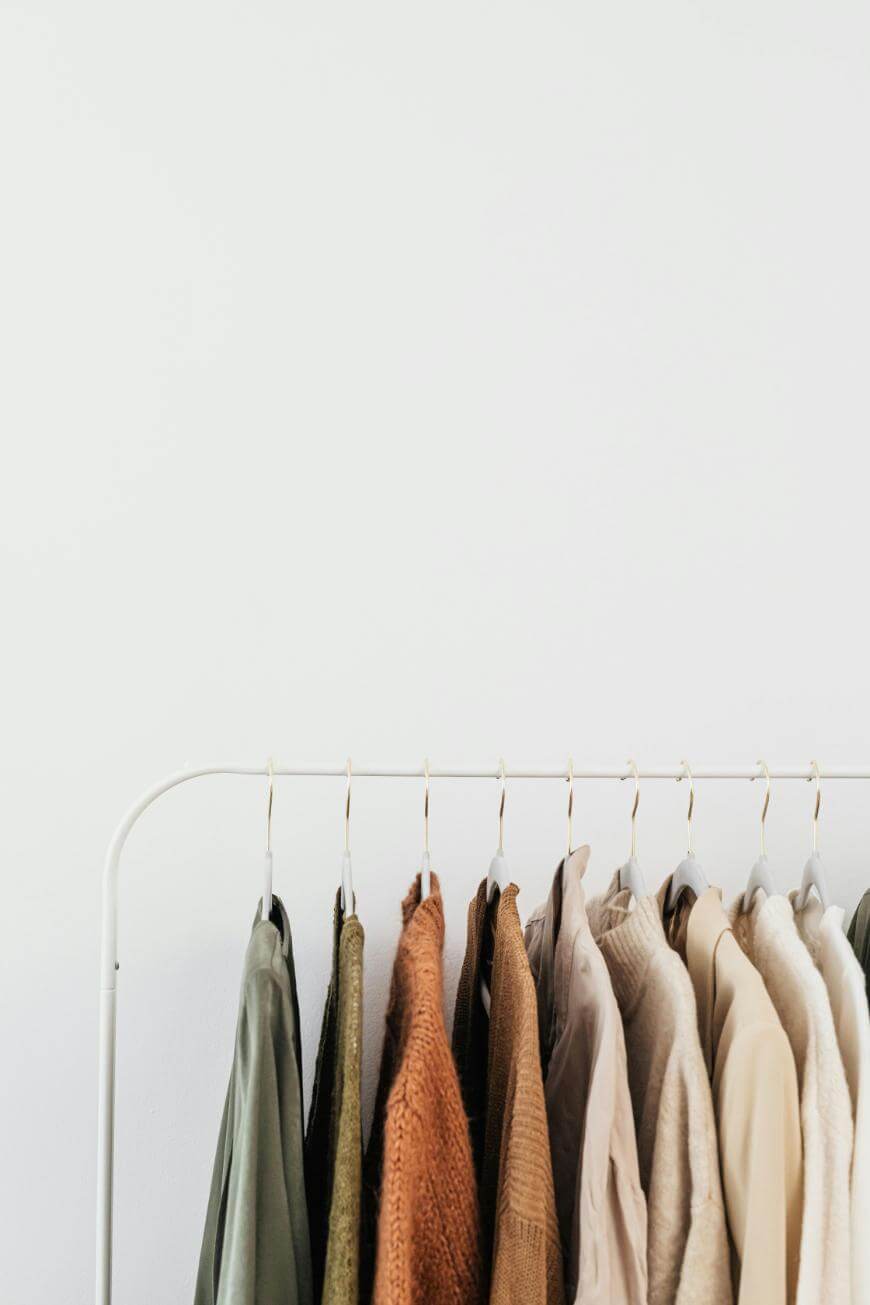 A minimalist clothing rack displays neatly hung garments in earthy tones, including greens, browns, and beiges, against a plain white wall.