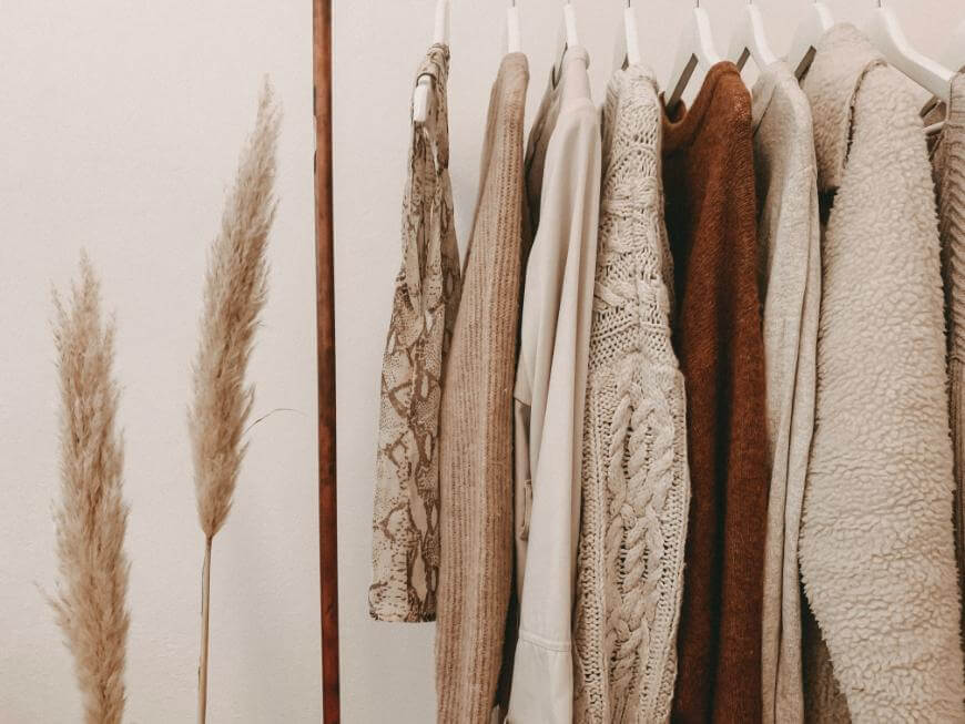 A clothing rack with various neutral-toned sweaters and knitwear on white hangers, next to tall dried pampas grass against a plain white wall.