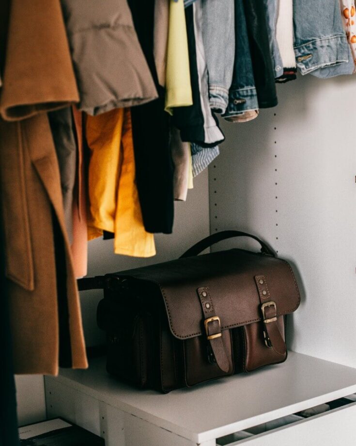 A brown leather satchel sits on a white shelf inside a closet, with various jackets, shirts, and pants hanging above it.