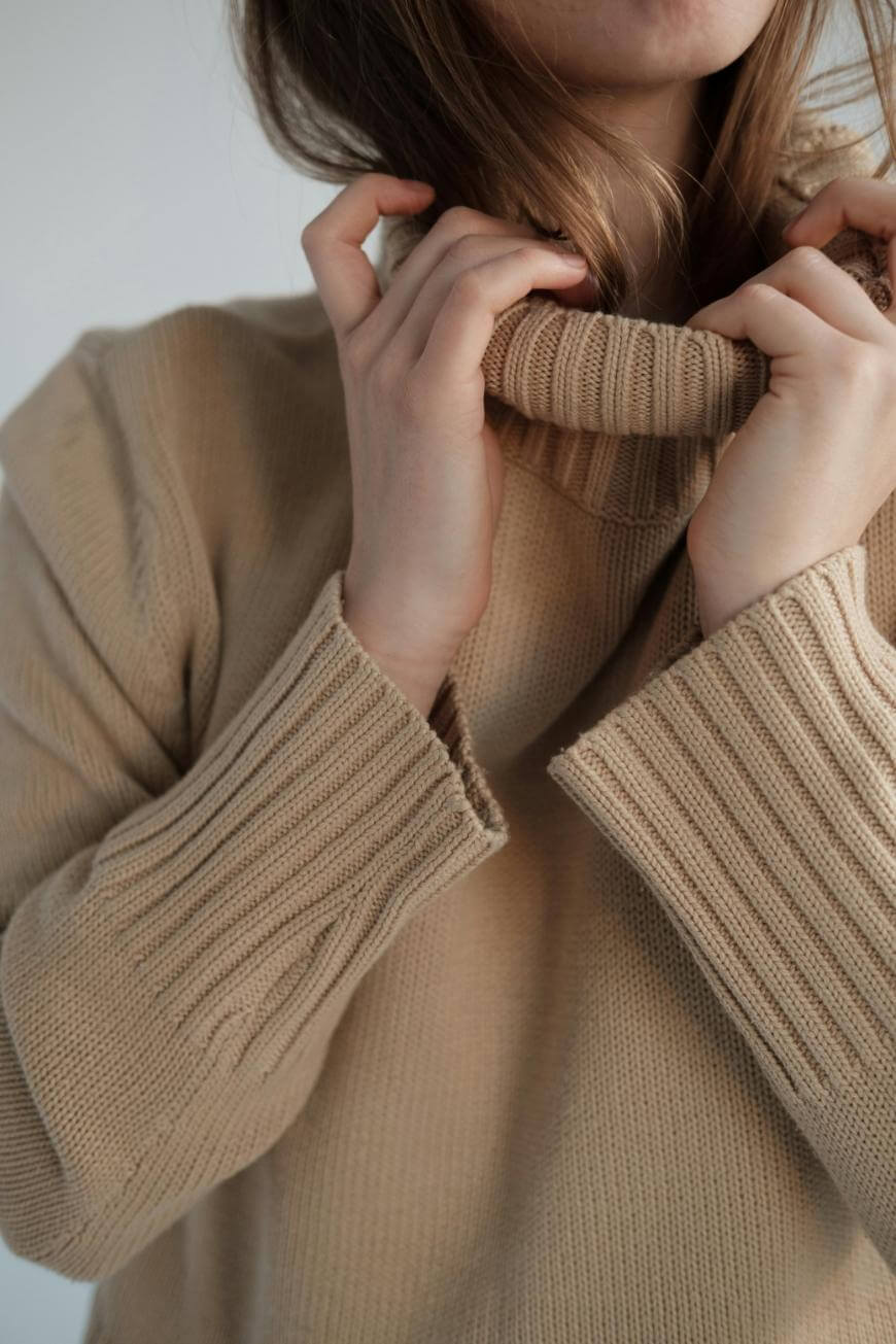 A person wearing a beige, ribbed knit sweater is gently pulling up the turtleneck collar, partially covering their face. The background is simple and out of focus.