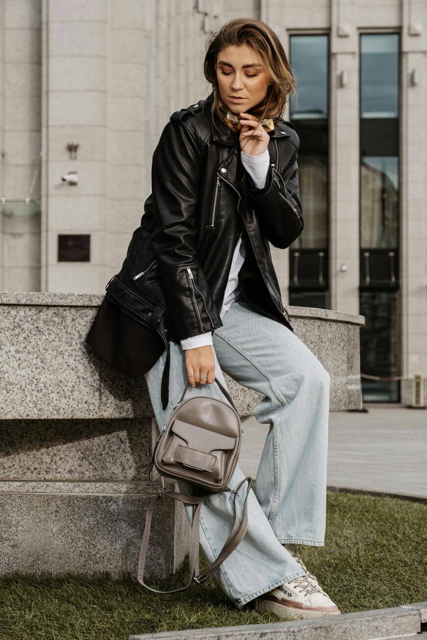 A woman wearing a black leather jacket, light blue jeans, and sneakers sits on a stone bench, holding a gray backpack and looking down while playing with her hair outside a modern building.