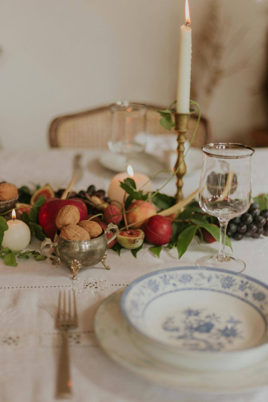 A table set with blue and white plates, a fork, and a wine glass. The centerpiece includes a candle, walnuts, grapes, figs, apples, and greenery on a white tablecloth.