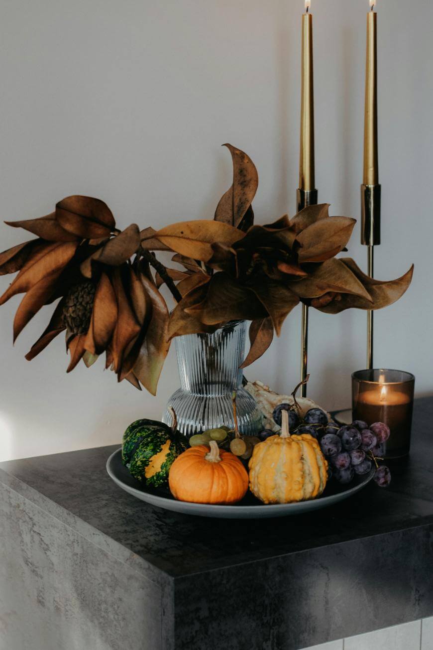 A decorative arrangement with dried brown leaves in a glass vase, two gold candlesticks, a lit candle, and a plate holding small pumpkins, gourds, and grapes on a dark surface.