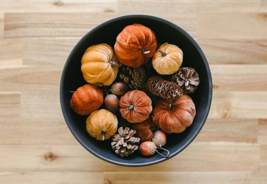 A black bowl filled with small decorative pumpkins, pine cones, and acorns sits on a light wooden surface. The pumpkins are orange and yellow, creating an autumn-themed arrangement.