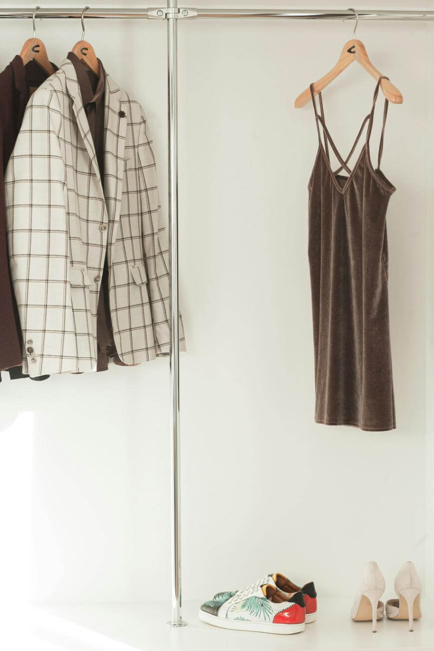 A minimalist wardrobe with a plaid blazer, dark jacket, strappy brown dress on hangers, and two pairs of shoes—colorful sneakers and beige high heels—arranged below on a white floor.
