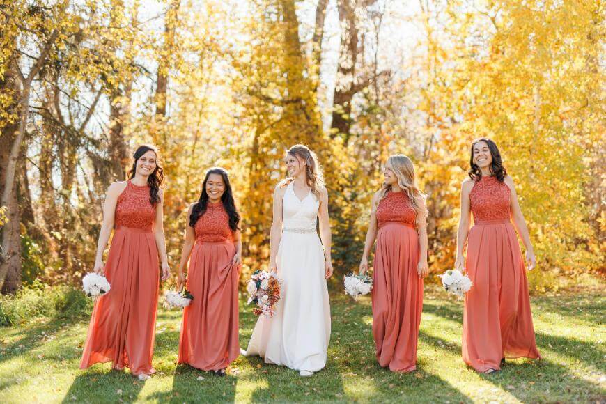 A bride in a white dress walks outdoors with four bridesmaids in matching rust orange dresses, all holding bouquets, surrounded by vibrant autumn trees with golden leaves.