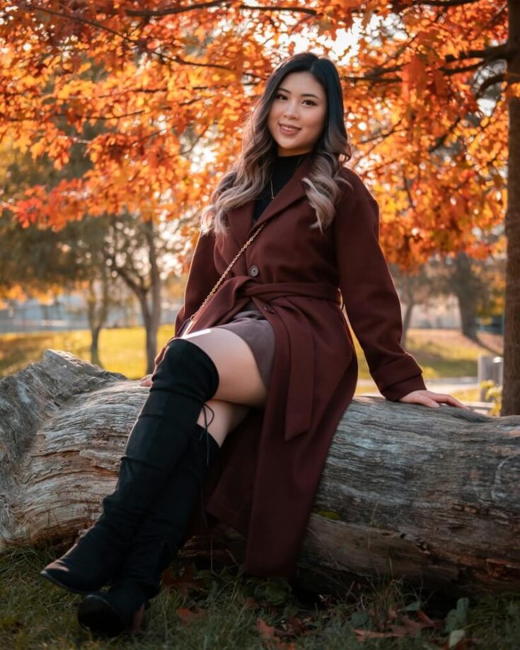 A woman with long hair sits on a large fallen tree trunk in a park, wearing a maroon coat and black knee-high boots. Autumn leaves in vibrant orange and red hues fill the background. She is smiling at the camera.
