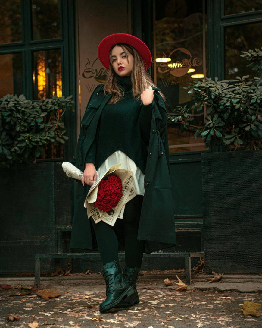 A woman in a black coat, black dress, and red hat stands outside a café holding a bouquet of red roses wrapped in newspaper. Leaves are scattered on the ground around her.