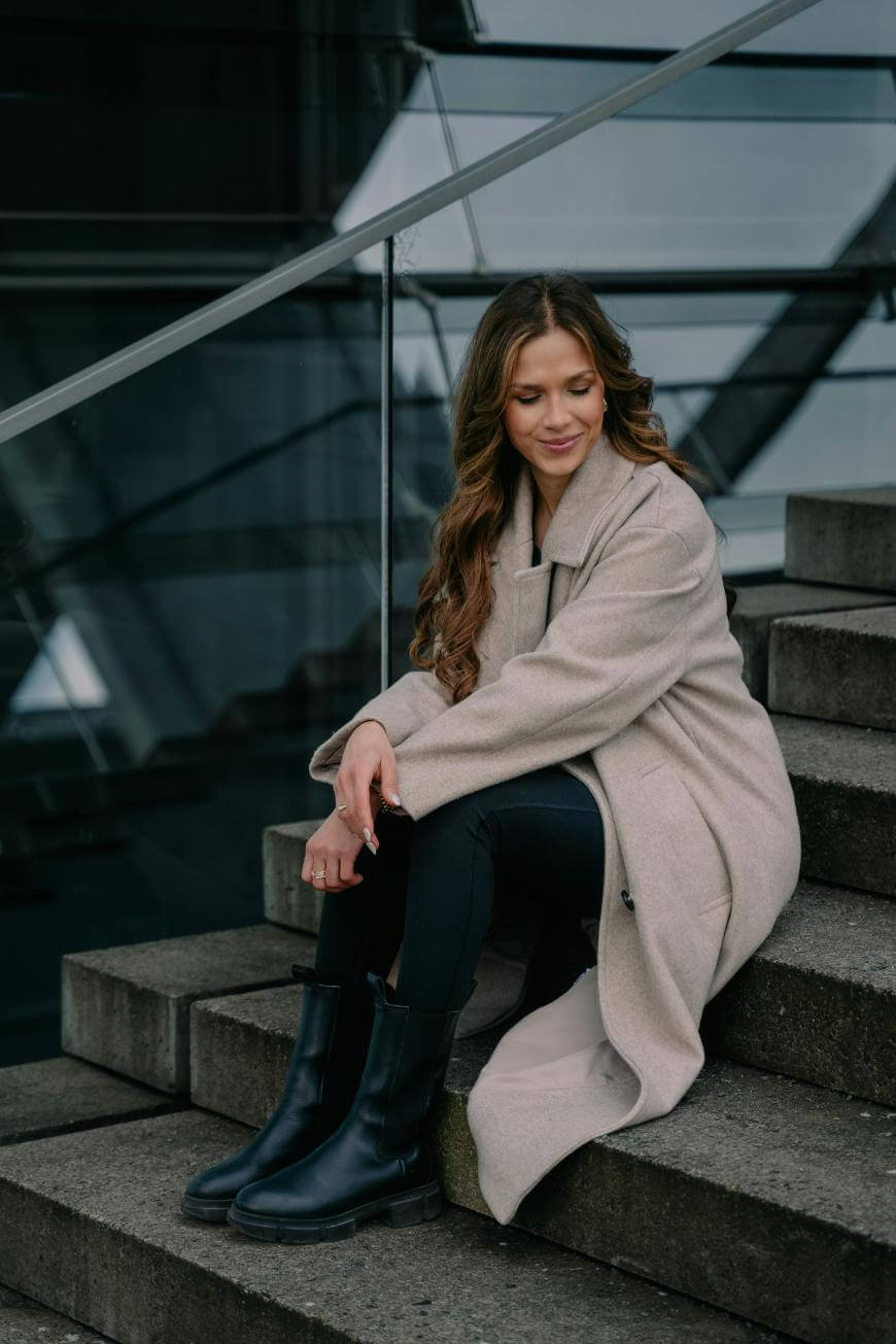 A woman with long wavy hair sits on outdoor concrete steps, wearing a light beige coat, black leggings, and black boots. She is looking down and smiling slightly, with a modern glass railing behind her.