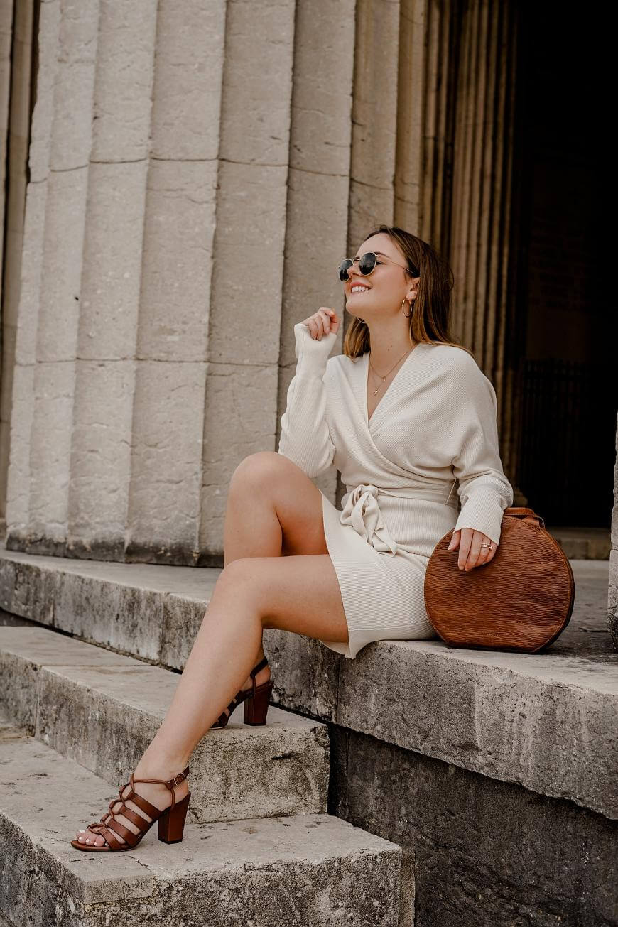 A woman in a cream-colored dress and brown heels sits on stone steps, holding a large round brown bag. She is smiling, wearing sunglasses, and appears relaxed near a columned building.