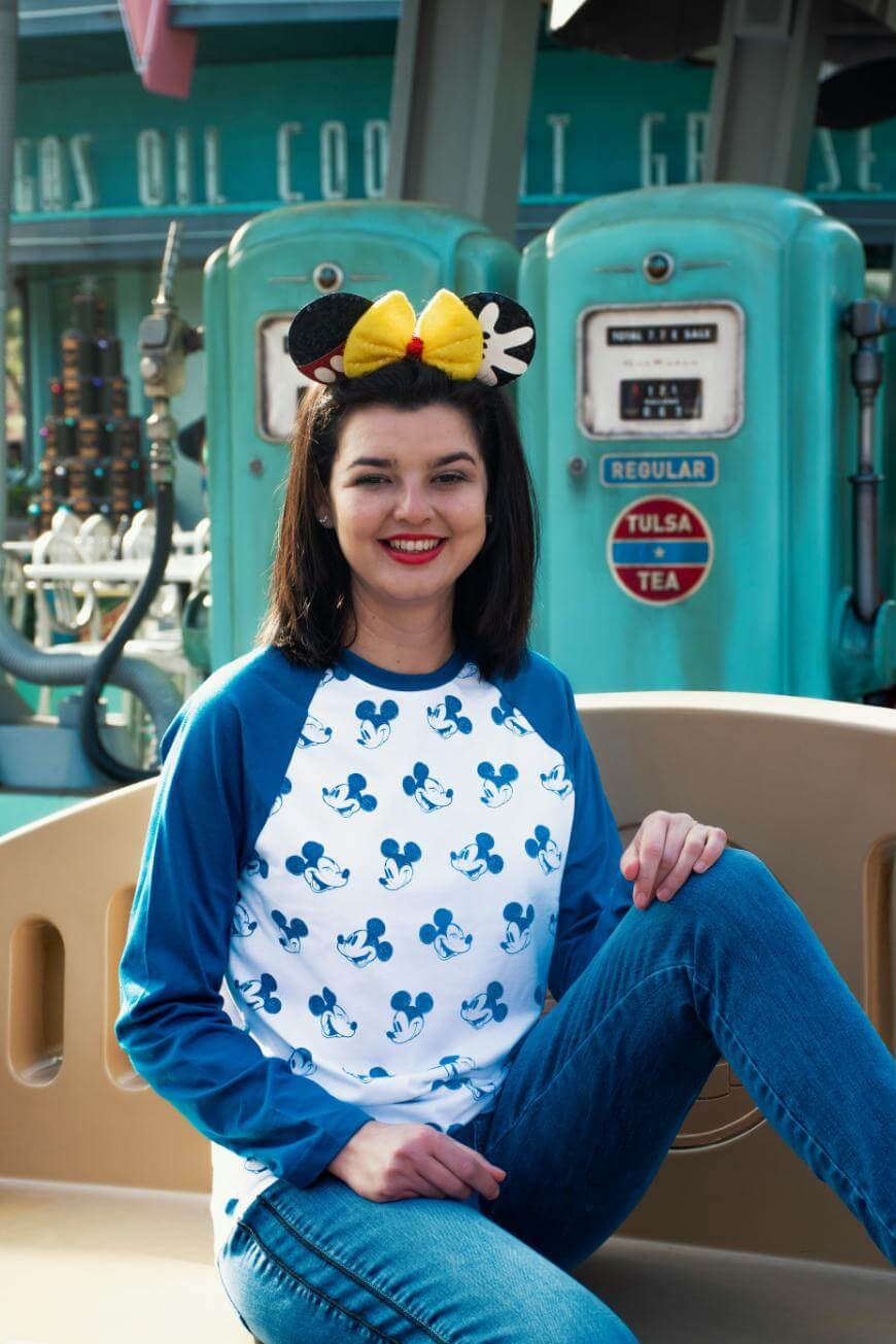 A smiling woman wearing a blue and white Mickey Mouse shirt, blue jeans, and Minnie Mouse ears sits in front of vintage teal gas pumps at a theme park.