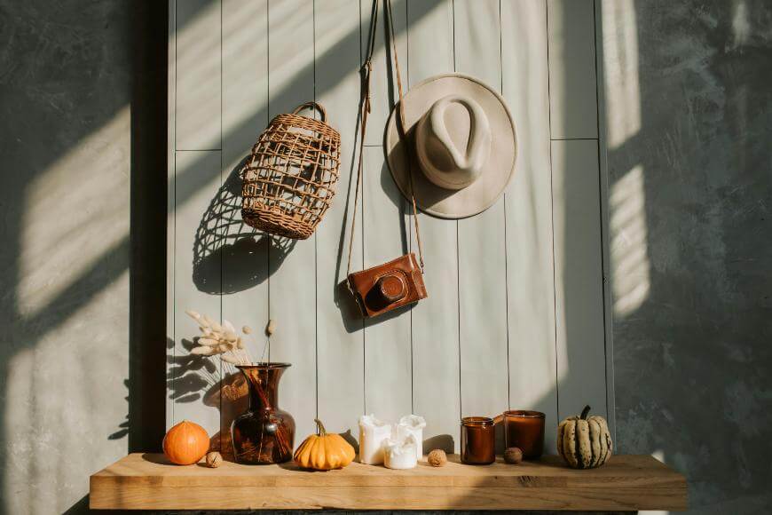 A wooden table holds pumpkins, candles, vases, and small decor. Above, a wicker bag, a hat, and a camera hang on a paneled wall. Sunlight casts shadows across the cozy autumn scene.