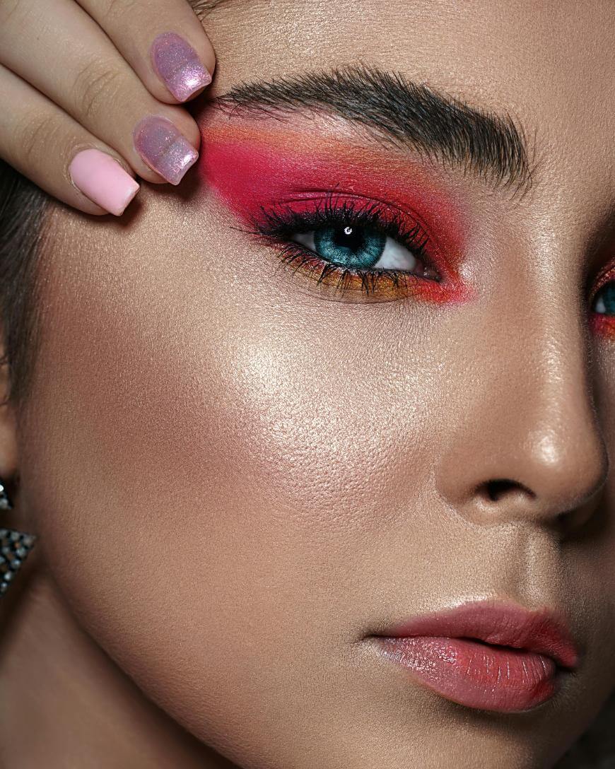 Close-up of a person with bold red and orange eyeshadow, dramatic lashes, groomed brows, glowing skin, nude lipstick, and manicured nails painted pink and purple, gently touching their forehead.
