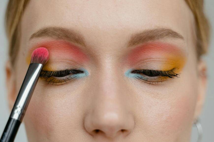 A close-up of a person applying pink eyeshadow to their eyelid with a makeup brush. Their eyelids are decorated with vibrant pink, yellow, and blue eyeshadow.