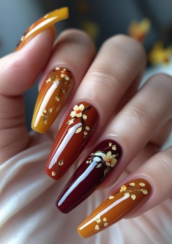 A hand with long, coffin-shaped nails painted in autumn shades of mustard yellow, burnt orange, and deep red, each decorated with delicate, small yellow floral designs.