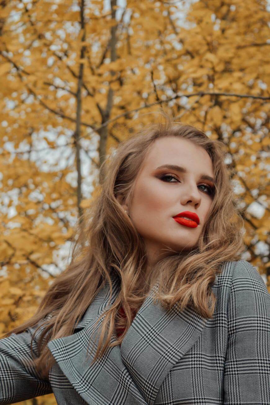 A woman with long, wavy blonde hair and bold red lipstick poses outdoors in front of trees with yellow autumn leaves. She is wearing a gray plaid coat and looking confidently at the camera.