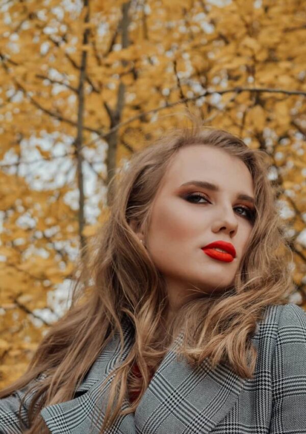 A woman with wavy blonde hair and bright red lipstick poses outdoors in front of trees with yellow autumn leaves, wearing a gray plaid blazer.