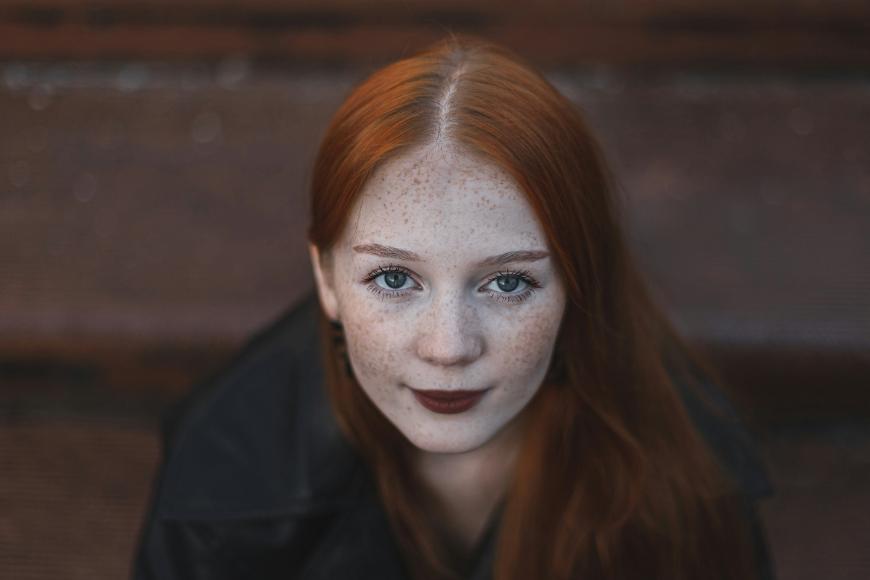 A young woman with long red hair, fair skin, and freckles looks up at the camera with a soft expression. She is wearing dark clothing and is seated in front of blurred brown wooden steps.
