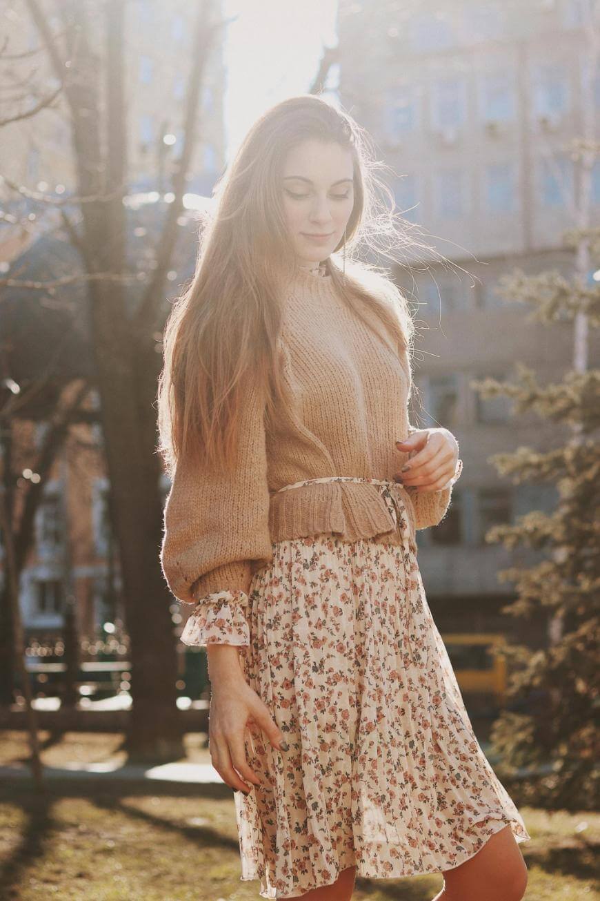 A woman with long hair stands outdoors in sunlight, wearing a light brown sweater over a floral dress. Trees and a building are visible in the background.