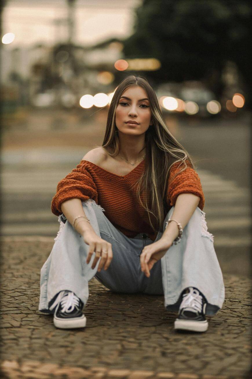 A young woman with long hair sits on a cobblestone street, wearing a rust-colored off-shoulder sweater, light ripped jeans, and black sneakers. The background is softly blurred with evening city lights.