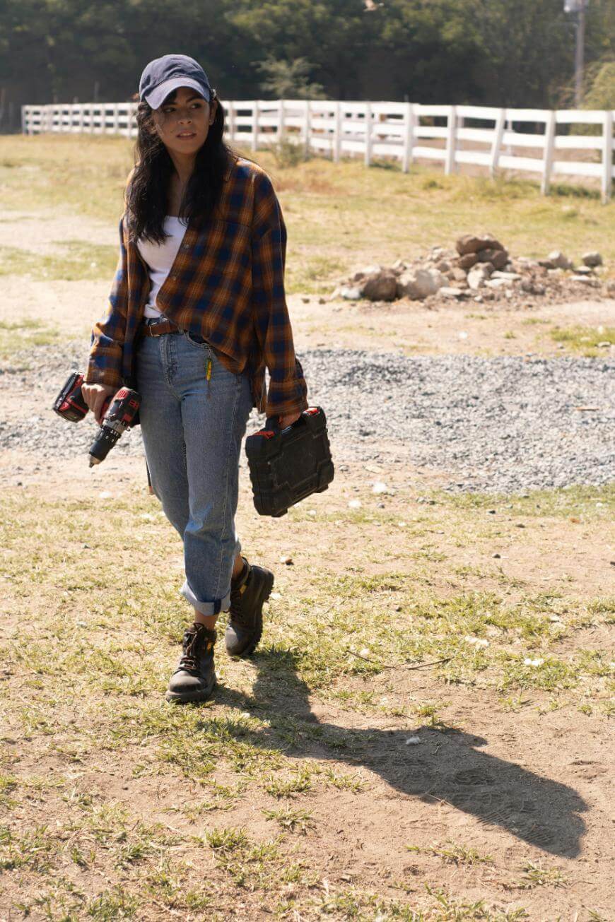 A woman in jeans, boots, a plaid shirt, and a cap walks outdoors on a sunny day, carrying a toolbox in one hand and a cordless drill in the other, with a white fence and trees in the background.