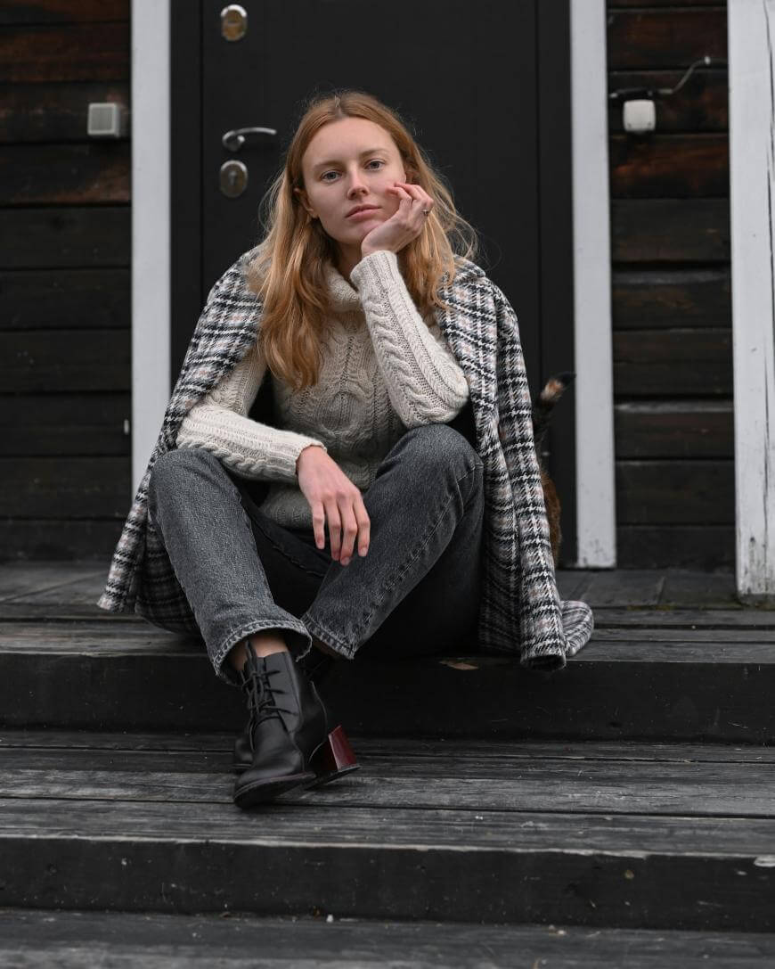 A young woman with long blonde hair sits on outdoor wooden steps, resting her chin on her hand. She wears a plaid coat, cream cable-knit sweater, gray jeans, and black boots, with a dark wooden door behind her.