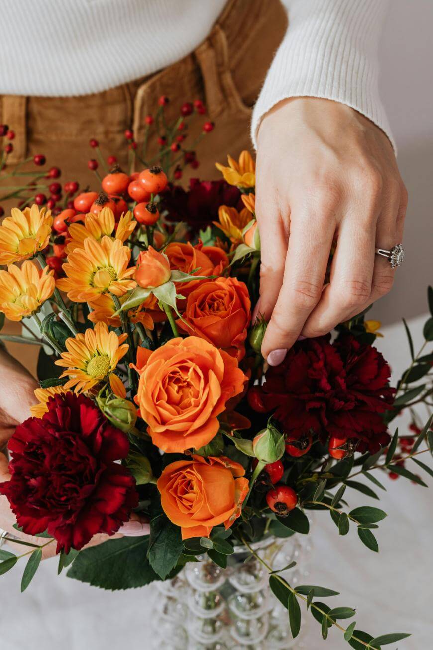 A person arranging a bouquet of orange roses, yellow chrysanthemums, and deep red carnations, wearing a white sweater and brown pants. A ring is visible on one hand.
