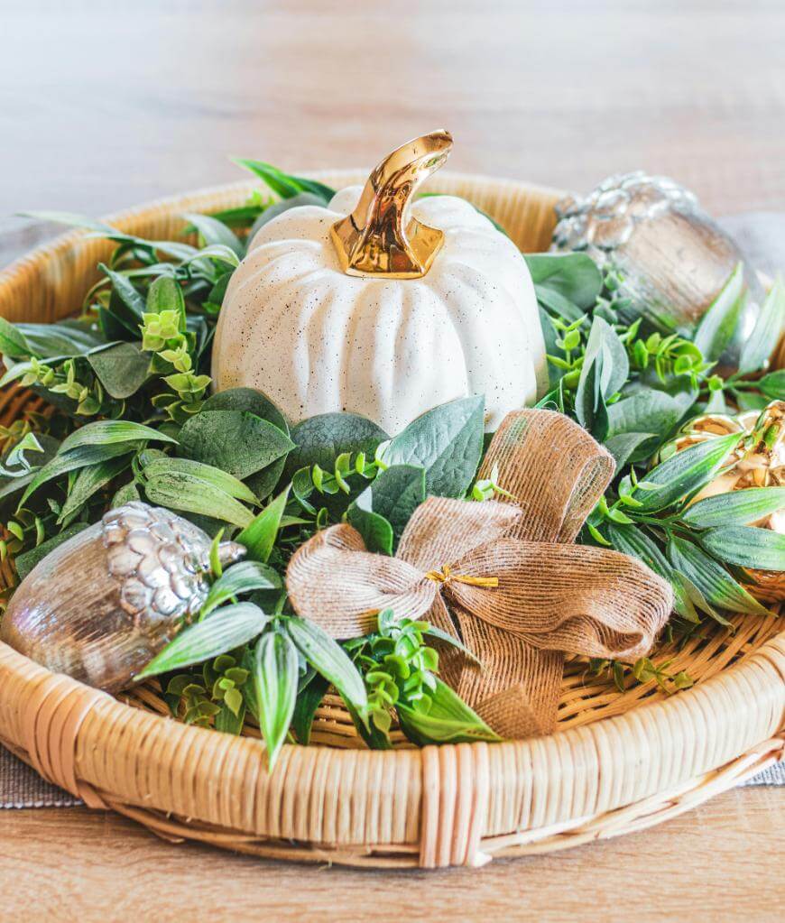 A woven tray holds a white pumpkin with a gold stem, surrounded by green foliage, metallic acorns, and a burlap ribbon, creating a festive autumn centerpiece.