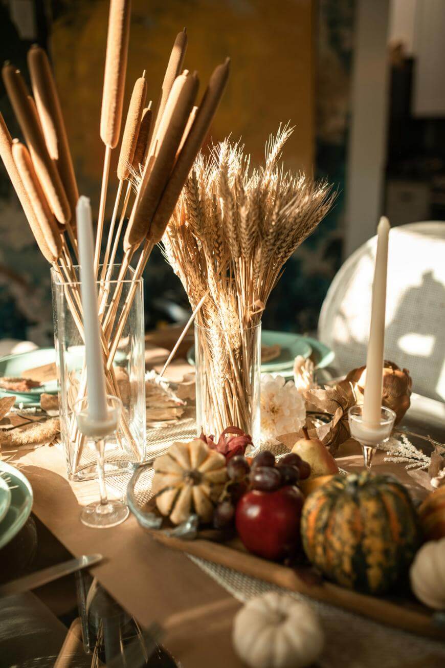 A festive autumn table setting features dried wheat, cattails in glass vases, white taper candles, and a centerpiece of assorted small gourds, grapes, and a pomegranate, all bathed in warm sunlight.