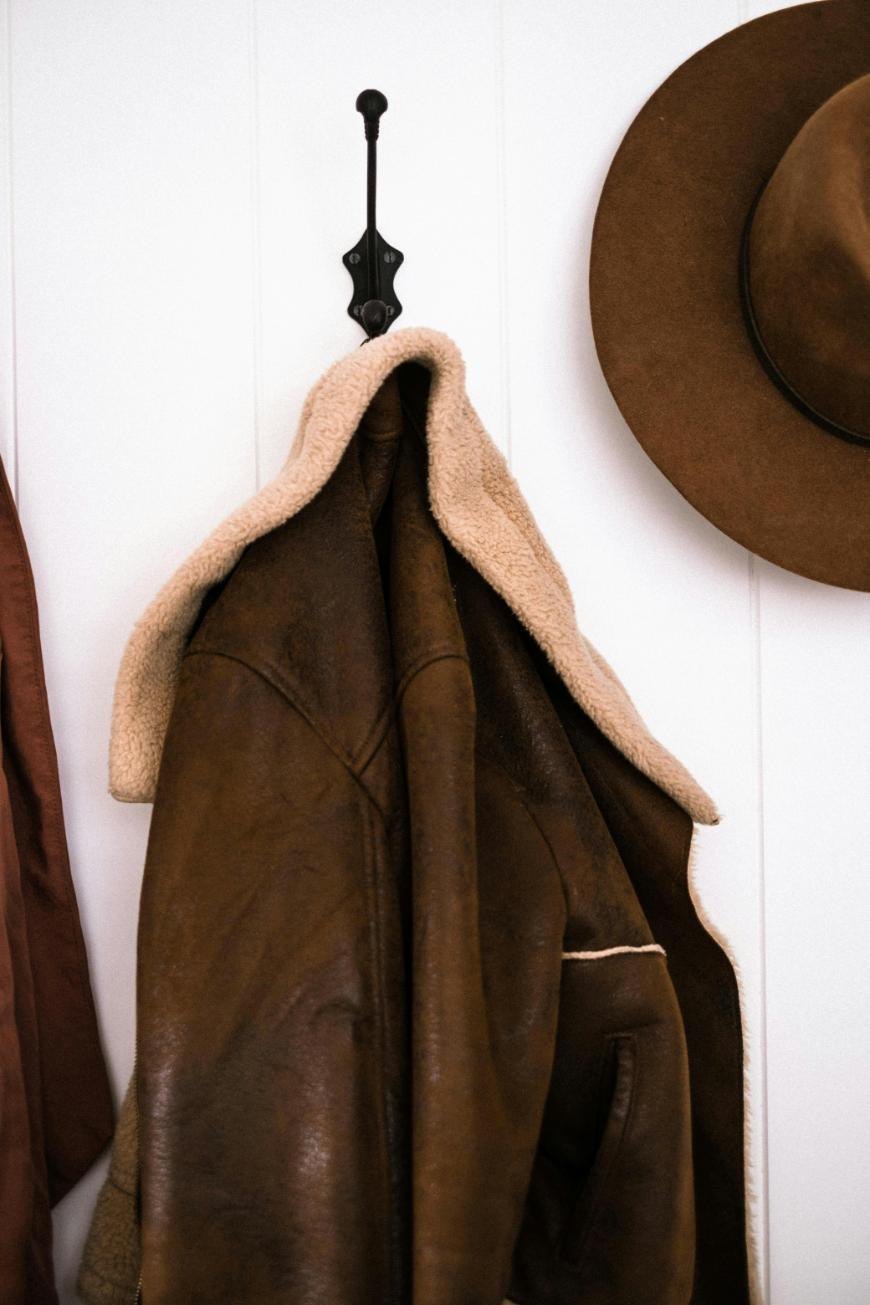 A brown shearling leather jacket hangs on a black metal wall hook, with a brown wide-brimmed hat hanging nearby on a white wall.