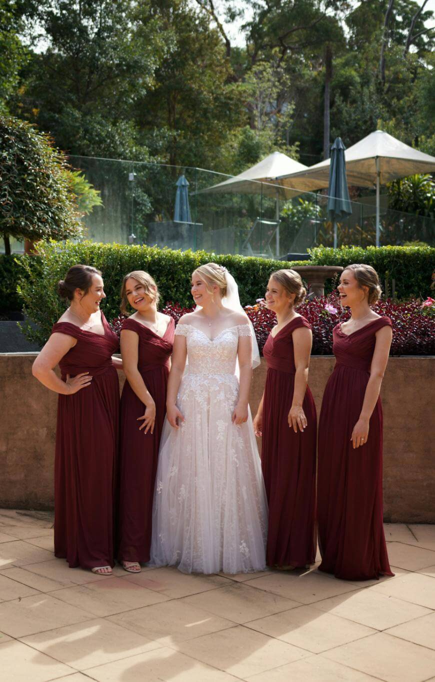 A bride in a white gown stands outside, smiling and surrounded by four bridesmaids in matching burgundy dresses. They are all laughing and posing together on a sunny day with trees and umbrellas in the background.