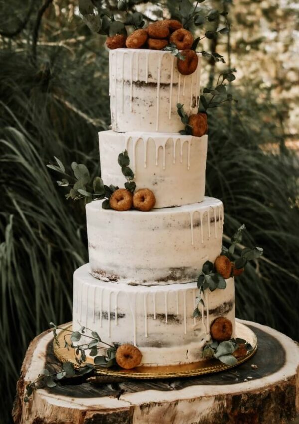 A four-tiered semi-naked cake with white icing drips, decorated with small donuts and green foliage, sits on a wooden slab outdoors with greenery in the background.