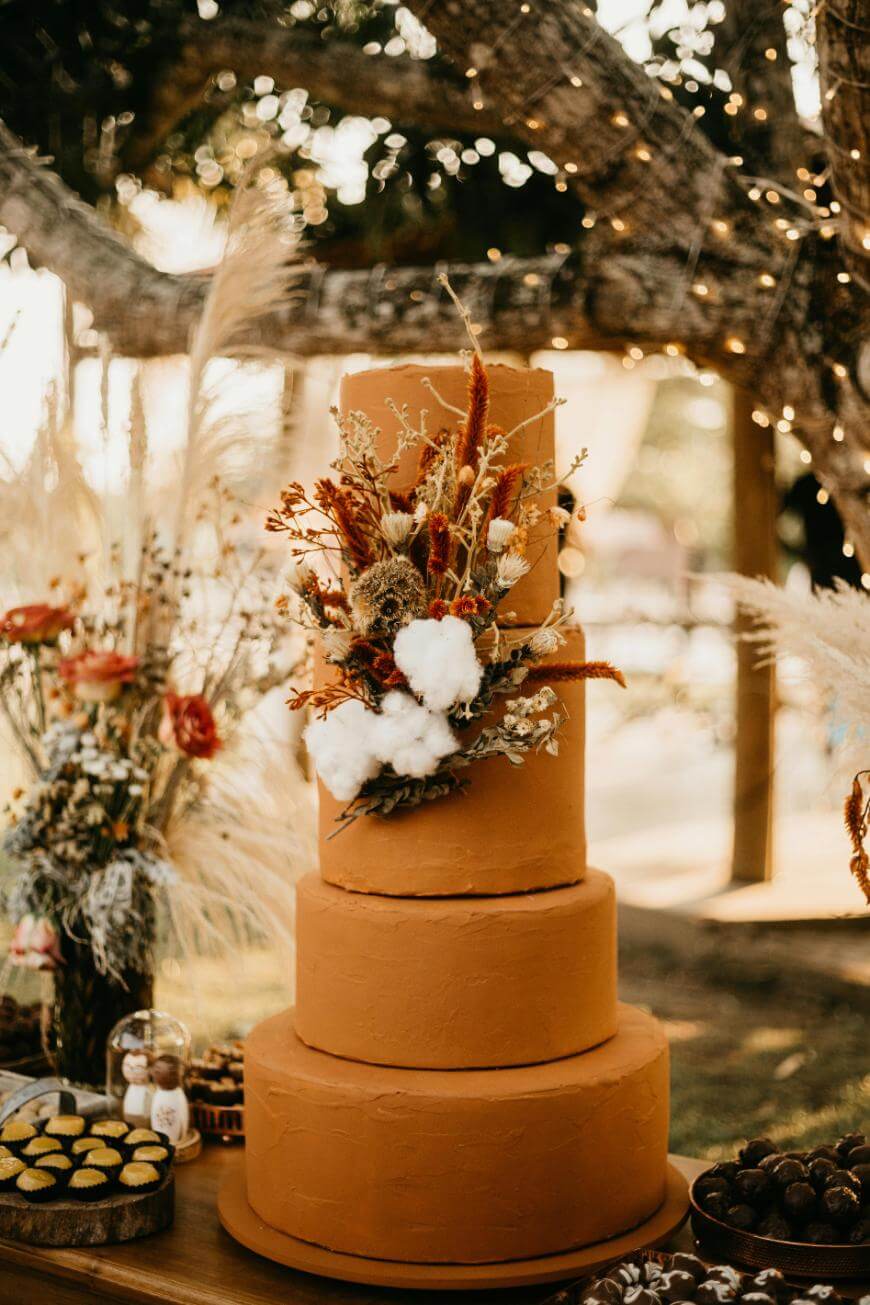 A four-tiered terracotta-colored wedding cake decorated with dried flowers, cotton, and grasses sits on an outdoor table under a tree with string lights, surrounded by rustic floral arrangements and desserts.